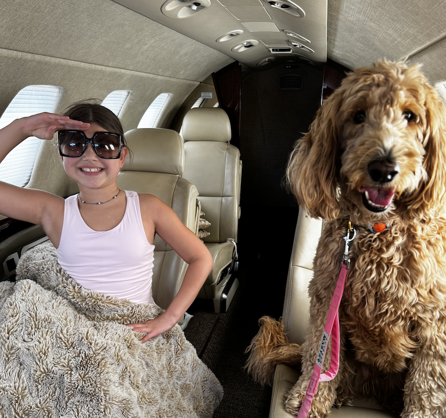 Izzy, a young girl with sunglasses saluting, covered by a fluffy blanket, sitting next to her dog on a private jet.